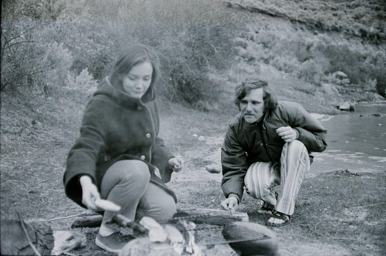 Two people crouched by a small fire roasting food on sticks